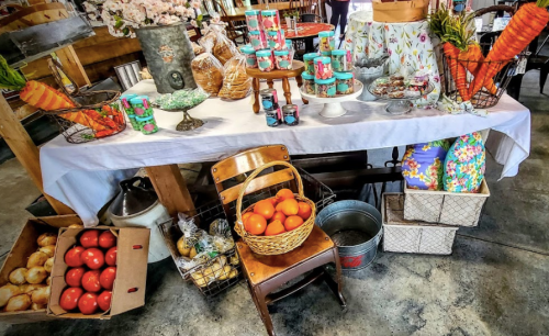 A rustic table display with colorful treats, flowers, and baskets of fruits, set in a charming market space.