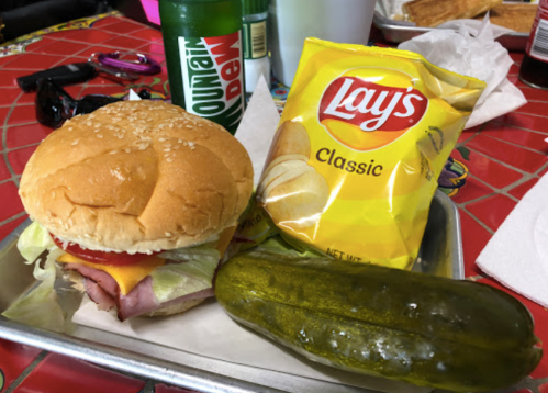 A sesame seed bun sandwich with ham, cheese, lettuce, tomato, a bag of Lay's chips, and a pickle on a tray.