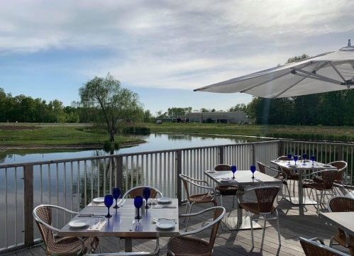 A scenic outdoor dining area with tables set by a calm pond, surrounded by greenery and a clear sky.