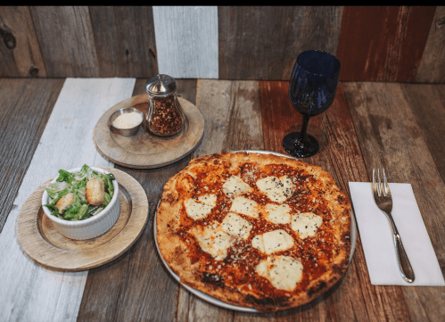 A wooden table with a pizza, salad, and a glass of wine, accompanied by condiments and a fork.