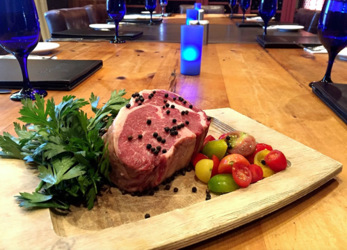 A raw ribeye steak garnished with black pepper, parsley, and colorful cherry tomatoes on a wooden platter.