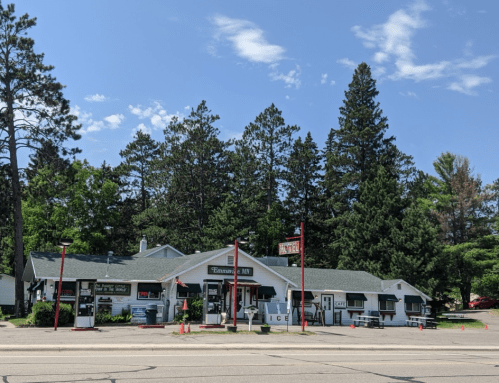 A small, charming store with a green roof, surrounded by trees, featuring signs for ice and a motel. Clear blue sky above.