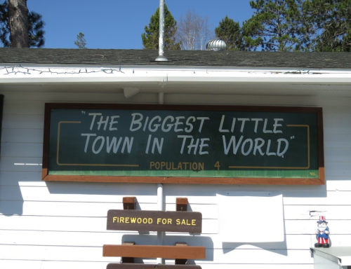 Sign reading "The Biggest Little Town in the World" with a population of 4, and a notice for firewood for sale.