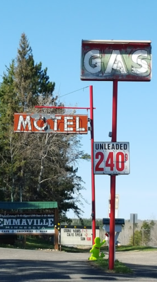A roadside scene featuring a vintage motel sign, a gas station sign, and a green figure near the road.