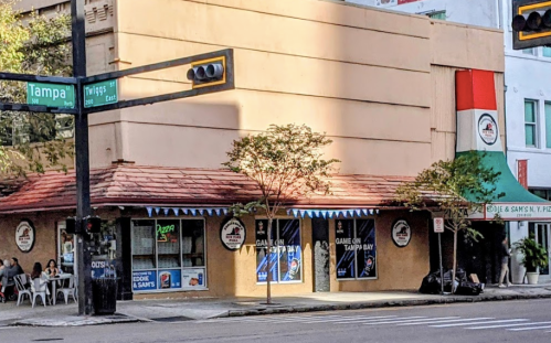 A street view of a restaurant with outdoor seating, featuring a red roof and colorful signage on a city corner.