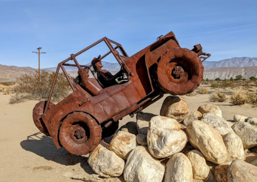 A rusty metal sculpture of a jeep climbing over a pile of rocks in a desert landscape.