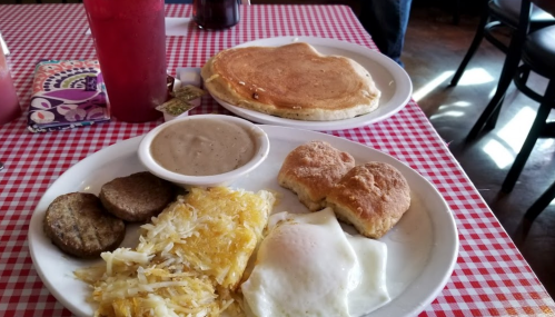 A breakfast plate with hash browns, sausage, a fried egg, biscuits, and gravy, alongside a pancake and a drink.