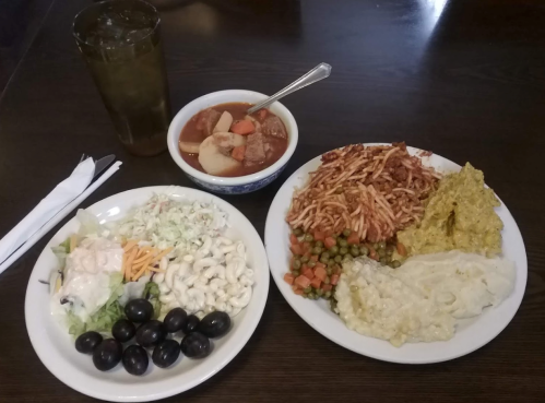 A plate of mixed foods including salad, pasta, vegetables, and a bowl of soup, with a drink on the side.