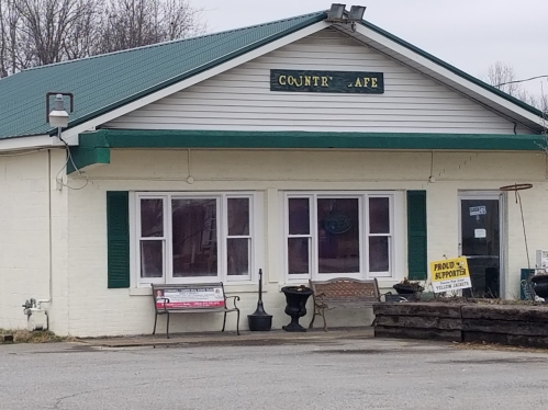 A small country café with a green roof, white walls, and large windows, featuring outdoor seating and a sign.