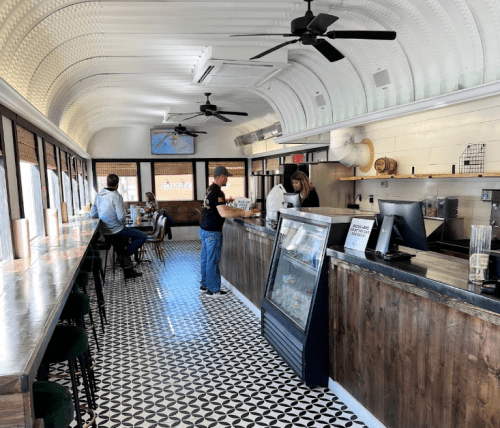 Interior of a modern café with a long counter, patrons ordering, and a patterned floor. Bright and inviting atmosphere.