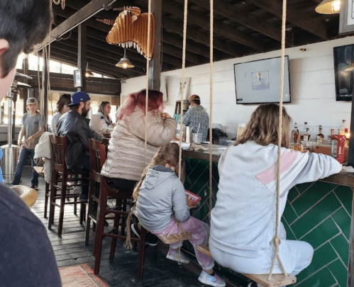 A cozy bar scene with patrons sitting on swings, enjoying drinks and conversation in a rustic setting.