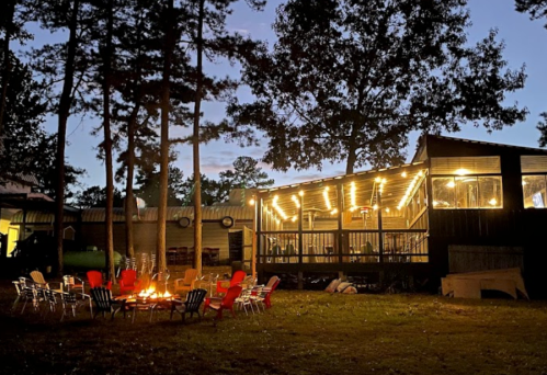 A cozy outdoor scene with a fire pit surrounded by colorful chairs, illuminated by string lights and trees in the background.