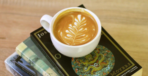 A white cup of latte art sits atop a stack of books on a wooden surface.