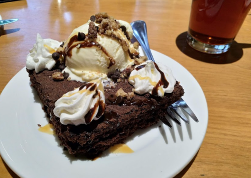 A slice of chocolate cake topped with ice cream, whipped cream, chocolate drizzle, and nuts, served on a white plate.