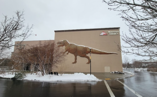 A large dinosaur mural on the side of a building, surrounded by snow-covered trees and a wet parking lot.