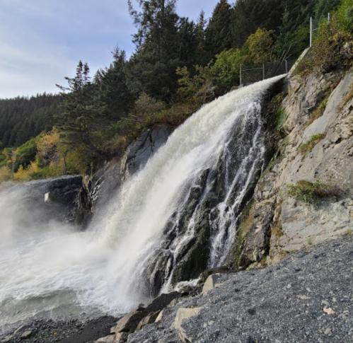 A waterfall cascading down rocky terrain, surrounded by trees and greenery under a clear sky.