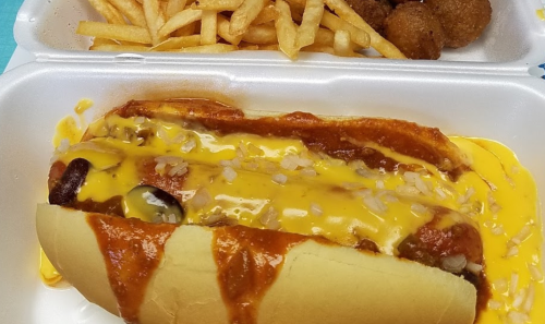 A hot dog topped with chili, cheese, and onions, served with fries and fried snacks in a takeout container.