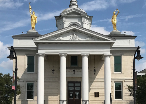 Historic building with golden statues atop columns, featuring a central entrance and blue sky in the background.