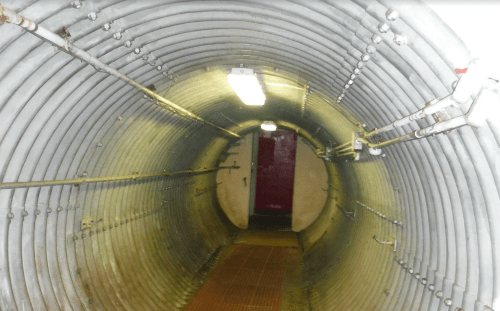 A dimly lit, cylindrical tunnel with metal walls and a red door at the end.