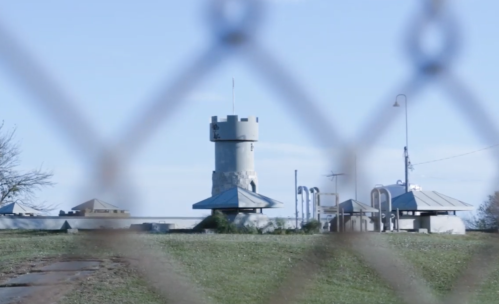 A stone tower and buildings are seen through a chain-link fence, with clear blue skies in the background.