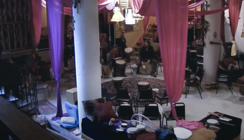 A cozy indoor space with drummers seated, colorful drapes hanging, and various instruments arranged around.