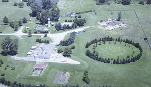 Aerial view of a rural property featuring a circular arrangement of trees and stone structures, surrounded by fields.