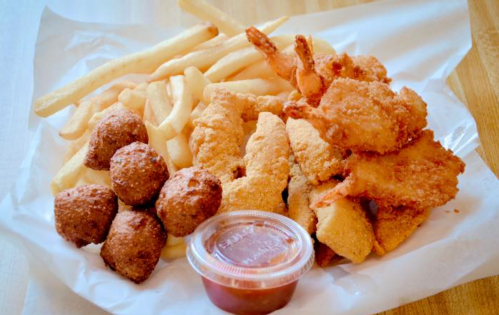 A plate of fried seafood including shrimp, fish fillets, hushpuppies, and French fries, served with a dipping sauce.