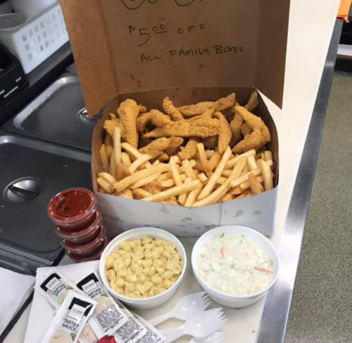 A large takeout box filled with chicken tenders and fries, accompanied by sides of macaroni, coleslaw, and dipping sauces.