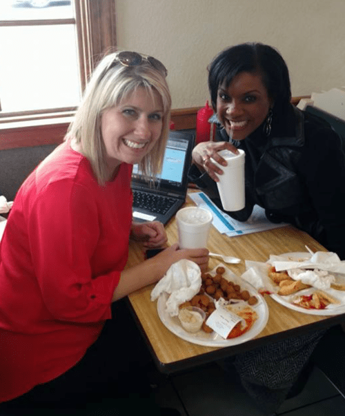 Two women smile at the camera while sitting at a table with food and drinks, a laptop in front of them.