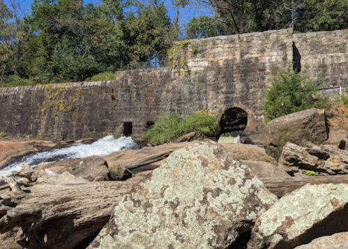 A rocky riverbank with a stone wall and a small water flow, surrounded by trees and greenery.