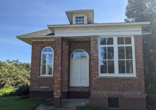 A brick building with large windows and a white door, set against a clear blue sky and greenery.