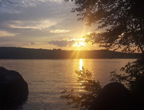 Sunset over a calm lake, with golden rays reflecting on the water and silhouettes of trees in the foreground.