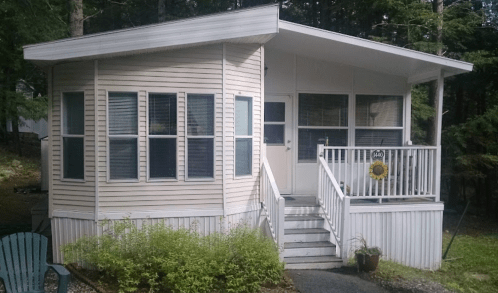 A small, light-colored house with a porch, surrounded by trees and greenery. Steps lead up to the front door.