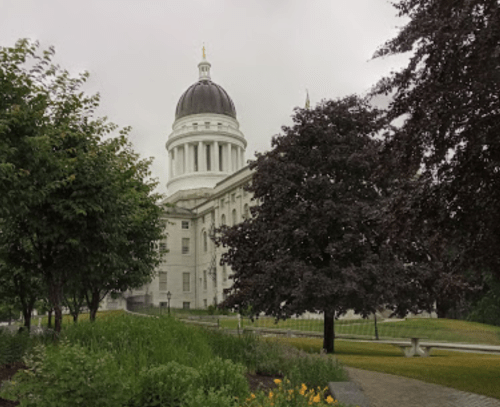 A view of a large white building with a dome, surrounded by green trees and a cloudy sky.