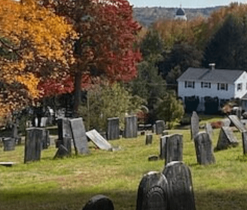 A peaceful cemetery with gravestones surrounded by colorful autumn trees and a distant house.