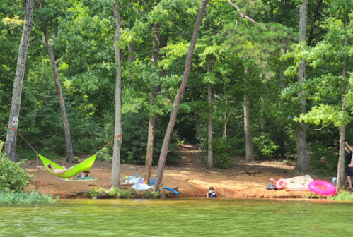 A serene lakeside scene with a green hammock between trees, surrounded by inflatable floats and a sandy shore.