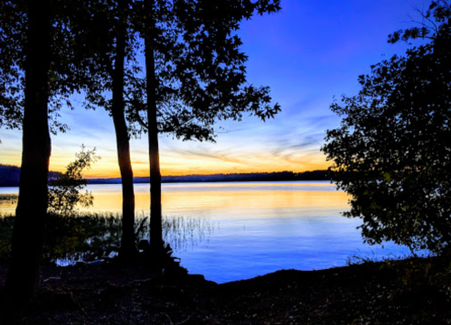 A serene lake at sunset, framed by trees, with vibrant blue and orange hues reflecting on the water's surface.