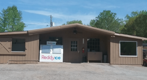 A brown wooden building with a sign for Reddy Ice, surrounded by trees and a clear blue sky.