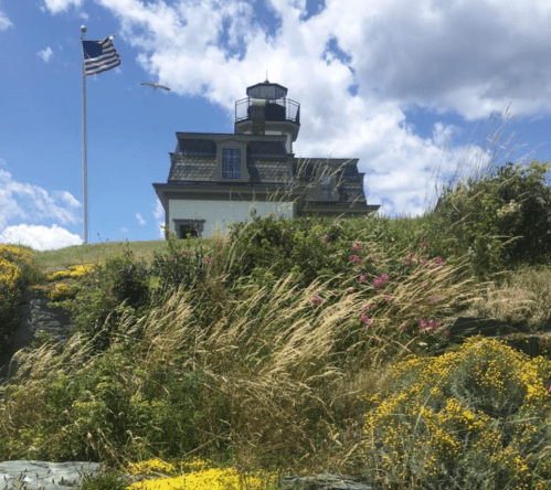 A lighthouse on a grassy hill with wildflowers, an American flag waving, and a cloudy blue sky above.