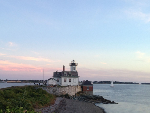 A lighthouse stands on a rocky shore at sunset, with a sailboat in the distance and soft pastel skies.