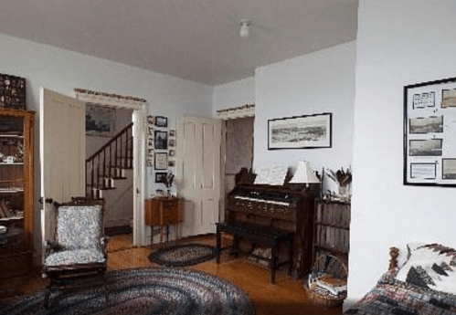 A cozy room featuring a piano, vintage furniture, and a staircase, with framed photos on the walls.