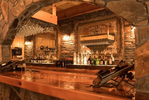 A rustic bar with a wooden counter, stone walls, and a variety of liquor bottles displayed on shelves.