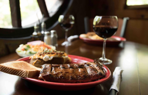A plate of grilled ribs with toast, a baked potato, and a side salad, accompanied by glasses of red wine on a wooden table.