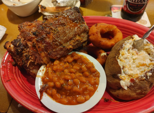 A red plate with BBQ ribs, baked beans, onion rings, and a loaded baked potato.