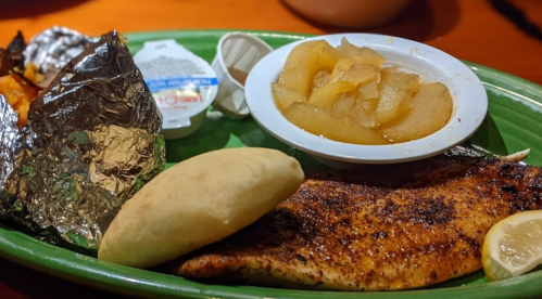 A plate featuring seasoned fish, a roll, baked potato wrapped in foil, and a side of sliced apples.