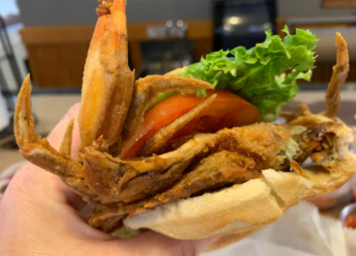 A hand holding a sandwich with a fried soft-shell crab, lettuce, and tomato, showcasing its crispy texture.