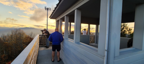 A person in a blue jacket stands on a balcony at sunset, while another person takes photos nearby.