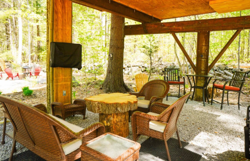 Cozy outdoor seating area with wicker chairs and a wooden table, surrounded by trees and a gravel floor.