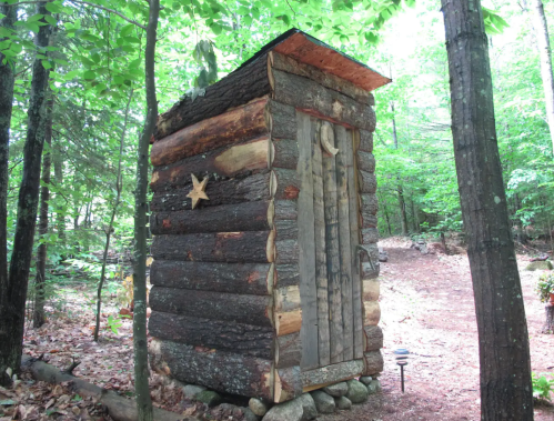 A rustic wooden outhouse surrounded by trees, featuring a star decoration and a natural stone base.