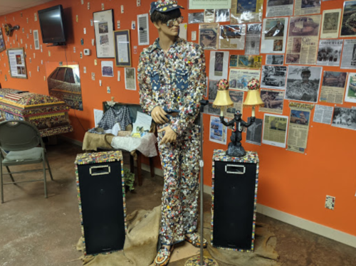 A mannequin dressed in a colorful, patterned outfit stands between two decorated speakers against a wall of newspaper clippings.